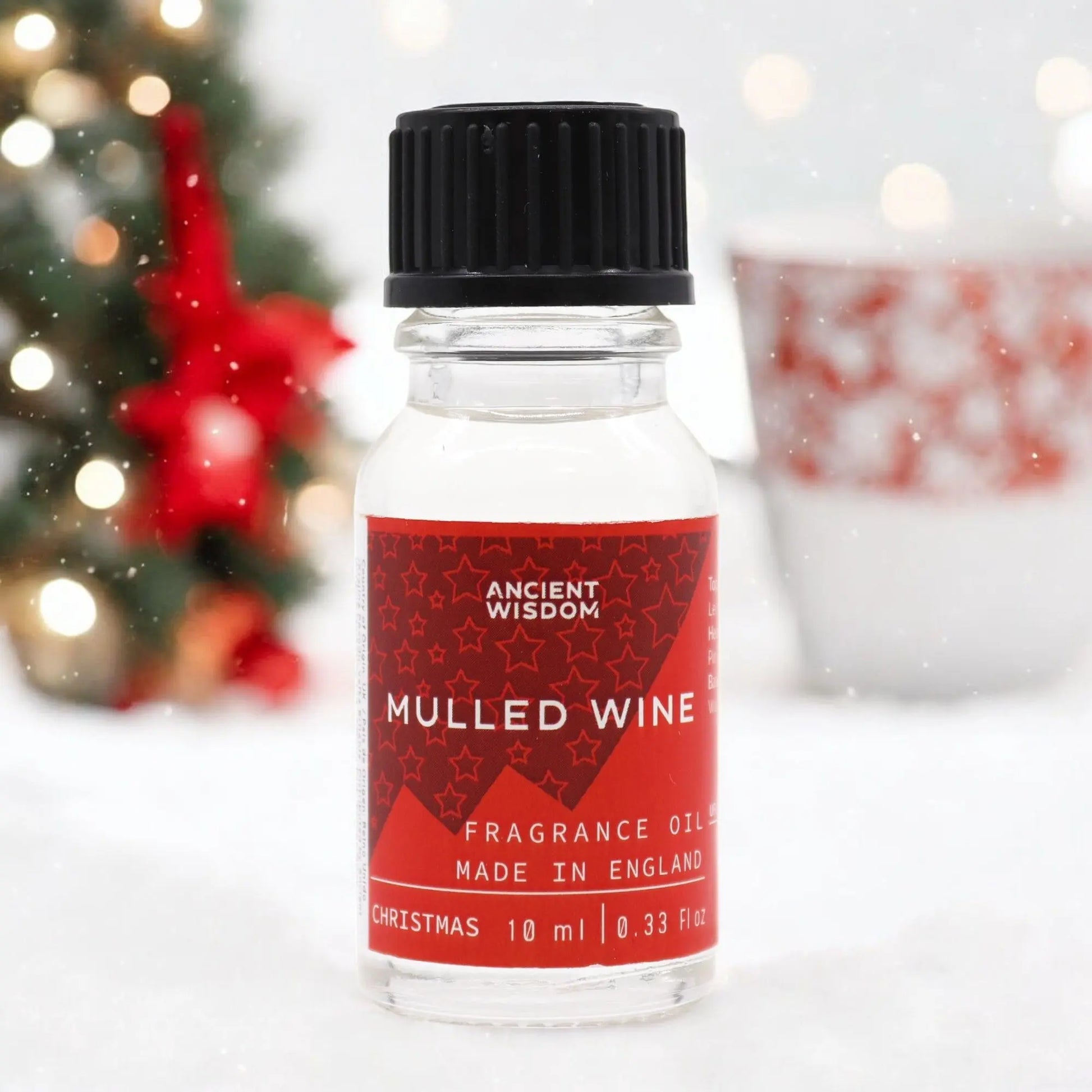 "Mulled Wine Fragrance Oil 10ml Ancient Wisdom in clear glass bottle with red star-patterned label, photographed against blurred Christmas tree with bokeh lights and red decorations"